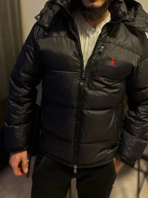 Svart pufferjacka från Polo Ralph Lauren - Snygg svart pufferjacka från Polo Ralph Lauren med huva och dragkedja framtill. Jackan har det klassiska röda Polo-logotypen på bröstet och flera praktiska fickor. Perfekt för kalla vinterdagar och riktigt skön att bära.