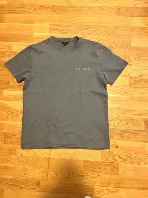Grå Essentials T-shirt Fear of God - Snygg grå T-shirt från Fear of God Essentials med diskret logga på bröstet. Klassisk rund halsringning och korta ärmar. Tillverkad i mjuk bomull som känns skön mot huden. Perfekt för en clean och stilren look.