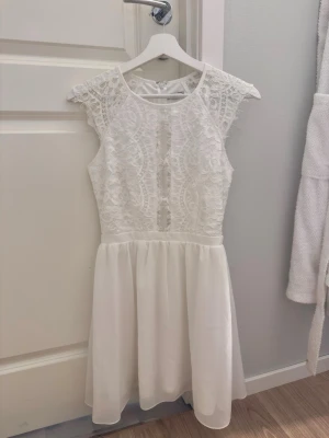 Nelly klänning - Säljer en söt vit, aldrig använd klänning från Nelly med spetsdetaljer upptill och en luftig kjol. Klänningen är ärmlös och har rund halsringning samt dragkedja i ryggen. Perfekt för studenten!