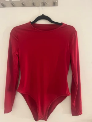 Röd långärmad body - Snygg röd body med långa ärmar och rund halsringning. Tillverkad i ett stretchigt, glansigt material som sitter tight mot kroppen. Perfekt att styla med jeans eller kjol för en trendig look.