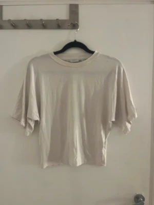 Beige croppad t-shirt med vida ärmar - En enkel och stilren vit t-shirt med croppad passform och vida, korta ärmar. Perfekt till jeans eller kjol för en avslappnad look. Tillverkad i mjuk bomull som känns skön mot huden. Den sitter åt k midjan och löst vid armarna