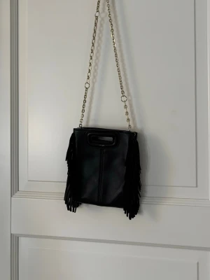  Maje handväska (äkta) - Cool svart handväska från maje med fransar på sidorna och två olika axelband – ett i guld kedja och ett i justerbart läder. Väskan har en fyrkantig form och ett praktiskt handtag upptill. Perfekt för dig som gillar  trendig stil. Den är dyr i nypris så säljer den för billigare 