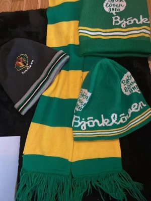 Grön och gul halsduk Björklöven - Säljer en klassisk halsduk från Björklöven i grönt och gult med breda ränder och fransar i ändarna och 3 mössor säljar allt i ett dom är väldigt gamla så det finns nån skada på halsduken och sen 2 små fläckar på ena mössan men dom är gamla 