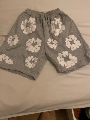 Grå shorts med vita blommor - Grå shorts med elastisk midja och vita blommönster över hela plagget. Tillverkade i mjukt bomullsmaterial för en avslappnad och bekväm känsla. Perfekta för varma dagar och har en loose passform.