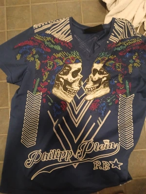 PP T-shirt  - t-shirt från Philipp Plein med stora dödskallemotiv framtill, färgglada detaljer och grafiska vita linjer. Baksidan har en stor geometrisk design och romerska siffror. Perfekt för dig som gillar statement-plagg och streetstyle.