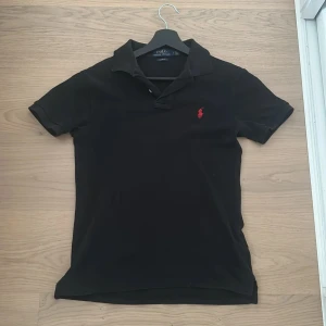Ralph lauren pike - Svart piké från Polo Ralph Lauren med klassisk krage och korta ärmar. Slim fit-modell med röd broderad logga på bröstet. Tillverkad i mjuk bomull som känns skön mot huden. Perfekt för dig som gillar stilrena och ikoniska plagg. Kom pv för eventuella frågor!😃