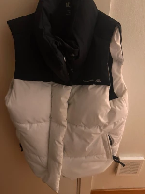 Svartvit dunväst med hög krage - Säljer en snygg svartvit dunväst med hög krage och dragkedja framtill. Västen har en puffig design, två sidofickor med dragkedja och justerbar nederkant. Perfekt för lager på lager och streetwear-stil.