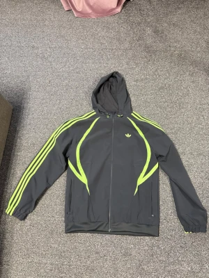 Adidas hoodie med neon detaljer - Mörkgrå hoodie från Adidas med dragkedja, huva och neon-gula detaljer längs ärmar och fram. Klassiska tre ränder på ärmarna och Adidas-logga på bröstet. Tillverkad i ett mjukt och sportigt material, perfekt för en streetwear-look.