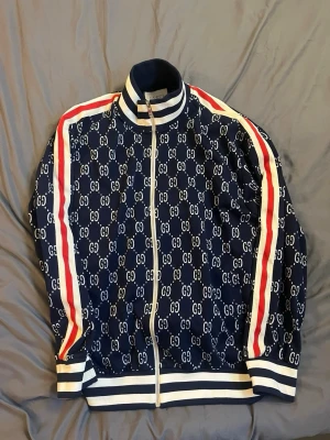 Marinblå Gcci zip med GG-mönster - Säljer en marinblå hoodie från Gci med helvitt G-mönster över hela plagget. Tröjan har vita och röda ränder längs ärmarna, dragkedja framtill och ribbade muddar i vitt och blått. Klassisk streetwear-look med hög krage och exklusiv känsla.
