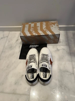 Golden Goose Superstar sneakers vit/röd - Golden Goose Superstar sneakers i vitt skinn med röd stjärna på sidan och svart häl. Grå snörning och platt sula i gummi. Klassisk rund tå och cool branding på plösen och hälen. Snygg kontrast mellan färgerna och detaljerna gör dem riktigt unika.