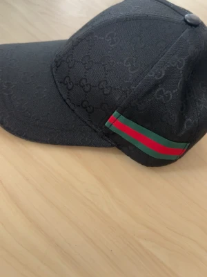Svart Gucci keps med GG-mönster - Säljer en svart keps från Gucci med klassiskt GG-mönster över hela kepsen och en grön-röd rand på sidan. Kepsen är tillverkad i bomull och har detaljer i polyester och elastan. Perfekt för dig som vill ha en stilren och lyxig accessoar.