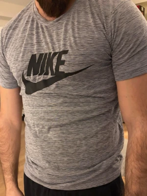Grå Nike t-shirt med logga - Säljer en gråmelerad t-shirt från Nike med stor svart logga på bröstet. T-shirten har rund halsringning och korta ärmar. Materialet känns mjukt och stretchigt, perfekt för en sportig look.
