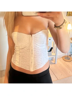 Vit broderad korsettopp - Supersnygg vit korsettopp med broderade detaljer och öppen axel. Toppen är axelbandslös och har en figurnära passform som framhäver midjan. Perfekt till jeans eller kjol för en trendig look.