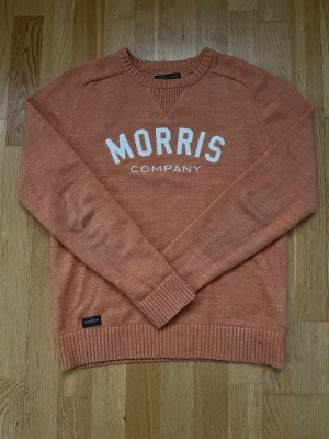 Orange tröja från Morris Gents - Snygg orange tröja från Morris Gents med vit text 'MORRIS COMPANY' på bröstet. Tröjan har rund halsringning, ribbade muddar och nederkant. Perfekt för dig som gillar stilrena och bekväma plagg med en sportig touch.
