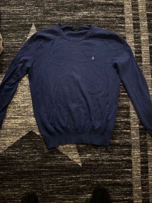 Mörkblå stickad tröja från Polo Ralph Lauren - Mörkblå stickad tröja från Polo Ralph Lauren med rund halsringning och klassisk broderad logga på bröstet. Tröjan har långa ärmar och ribbade muddar vid ärmslut och nederkant. Perfekt för en stilren och avslappnad look. ÄKTA SÅKLART