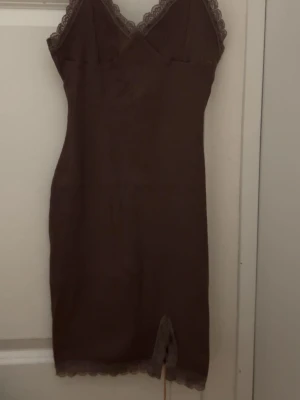 Brun klänning  med spetsdetaljer - Snyggt brunt nattlinne med smala axelband och spetsdetaljer vid urringning och nederkant. Figurnära modell med liten slits framtill. Perfekt för mysiga kvällar hemma.