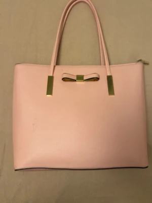 Ljusrosa handväska med rosett - Söt ljusrosa handväska i slät konstläder med två handtag och dekorativ rosett i guld på framsidan. Väskan har guldfärgade detaljer och dragkedja upptill. Perfekt för dig som gillar stilrena accessoarer med en feminin touch.