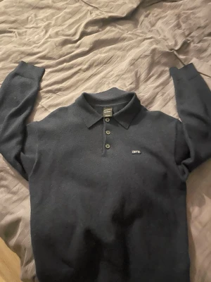 Mörkblå stickad tröja från Iets Frans - Mörkblå stickad tröja från Iets Frans med klassisk krage och tre knappar framtill. Tröjan har lång ärm och en liten broderad logga på bröstet. Perfekt för dig som gillar en stilren och enkel look. Köpt i barcelona för övrigt