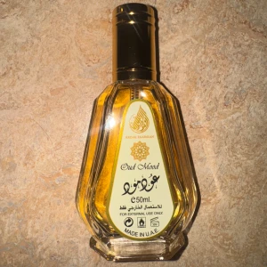 Oud Mood parfym 50ml från Ard Al Zaafaran - Oud Mood parfym från Ard Al Zaafaran i en elegant, sexkantig glasflaska med guldfärgad vätska och svart/guldfärgad kork. Flaskan rymmer 50 ml och har detaljerad etikett med arabiska och engelska texter. Tillverkad i Förenade Arabemiraten.
