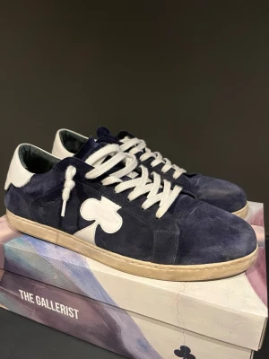 Mörkblå sneakers från The Gallerist - Snygga mörkblå sneakers i mocka från The Gallerist med vita detaljer och klubb-symbol på sidan. Skorna har vita skosnören och vit hälkappa, samt en beige sula. Perfekta för dig som gillar unik design och streetstyle.