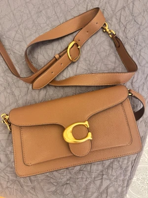Brun axelväska från Coach - Snygg brun axelväska från Coach i slätt läder med justerbar axelrem och guldfärgade metalldetaljer. Väskan har ett stort C-spänne framtill och en praktisk ficka på baksidan. Perfekt för dig som gillar stilrena accessoarer med lyxig känsla.