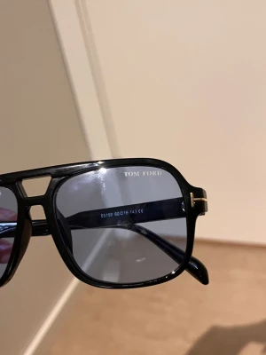 Svarta solglasögon från Tom Ford - Snygga svarta solglasögon från Tom Ford med fyrkantig form och mörka glas. Bågarna har en diskret gulddetalj vid gångjärnen och klassisk logga på sidan. Tillverkade i Italien och perfekta för en trendig look.