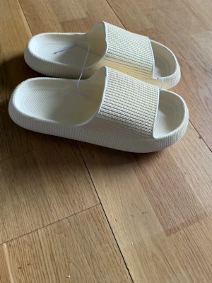 Beige Tofflor med ribbad rem - Säljer ett par beige tofflor med bred ribbad rem och tjock sula. Tillverkade i mjukt och flexibelt gummimaterial för extra komfort. Perfekta för en avslappnad och trendig look. Tofflorna är i storlek 36-38