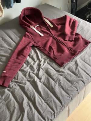 Vinröd russemerchhoodie med dragkedja - Säljer en vinröd russemerchhoodie med dragkedja och vita snören i huvan. Hoodien har två fickor framtill och ribbade muddar vid ärmar och nederkant. Perfekt för en avslappnad och chill stil.