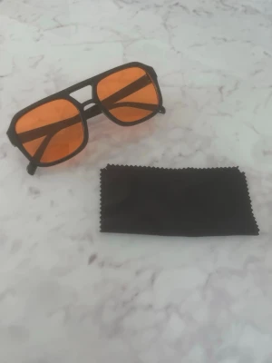 Svarta solglasögon med orange glas - Snygga solglasögon med svart båge och rektangulär form. Glasen är orangefärgade och ger en cool retrovibe. Perfekta för att sticka ut i sommar. Rengöringsduk i svart medföljer. Helt nya och oanvända!