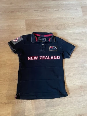  New Zealand skjorta East West - Svart t-shirt från East West med 'NEW ZEALAND' tryck på bröstet och flagga. Stor siffra 3 på ryggen och detaljer på ärmen. Klassisk pikékrage och korta ärmar. Tillverkad i bomull för skön känsla.                                                                        Pris kan diskuteras vid snabb affär