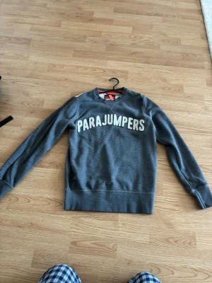 Grå sweatshirt från Parajumpers - Snygg grå sweatshirt från Parajumpers med stor vit logga på bröstet. Tröjan har rund hals, långa ärmar och ribbade muddar. Detaljer som orange band bak och patch på ärmen ger extra stilpoäng. Perfekt till jeans eller joggers.