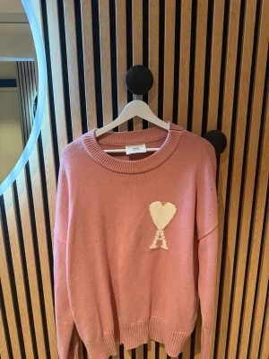 Rosa stickad tröja från Ami - Säljer en snygg rosa stickad tröja från Ami Alexandre Mattiussi med ett vitt hjärta och bokstaven A broderat på bröstet. Tröjan har rund halsringning och ribbade muddar. Perfekt för dig som gillar trendiga och bekväma plagg.