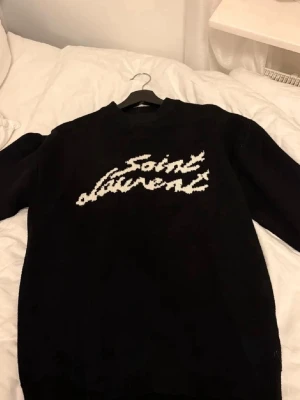 Svart stickad Saint Laurent-tröja - Svart stickad tröja från Saint Laurent med vit broderad logga framtill i coolt, handskrivet typsnitt. Tröjan har rund halsringning och långa ärmar. Perfekt för dig som gillar streetstyle och exklusiva märken.