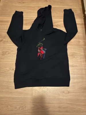 Svart hoodie från Polo Ralph Lauren - Snygg svart hoodie från Polo Ralph Lauren med stor broderad polospelare i färgglada nyanser på bröstet. Klassisk huva och ribbade muddar. Perfekt för en avslappnad och trendig stil.