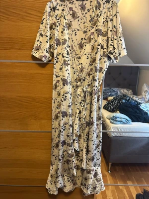Blommig långklänning från Soaked - Långklänning från Soaked med blommigt mönster i beige, svart och lila toner. Omlottmodell med v-ringning, knyt i midjan och volangkant nertill. Halvlånga ärmar och lätt, luftig känsla. Perfekt för sommarens alla tillfällen.