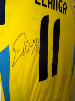 Sverige matchtröja Elanga #11 signerad - Gul och blå matchtröja från Sveriges landslag med Elanga och nummer 11 på ryggen. Tröjan är signerad och har svarta detaljer samt meshpaneler på sidorna för ventilation. Perfekt för fotbollsfans och samlare!