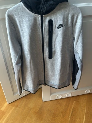 Grå Nike hoodie med svarta detaljer - Snygg grå hoodie från Nike med svarta detaljer och dragkedja framtill. Har en svart huva och en vertikal bröstficka med dragkedja. Tillverkad i mjukt material, perfekt för en sportig och avslappnad stil. Nike-logga på bröstet för extra streetkänsla.