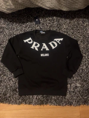Svart PRADA Milano hoodie - Snygg svart hoodie från PRADA med stort vitt PRADA-tryck över bröstet och texten MILANO under. Klassisk passform med ribbade muddar vid ärmar och midja. Perfekt för dig som vill ha en stilren och exklusiv look.
