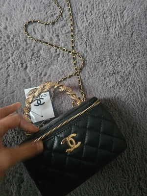 Svart quiltad handväska - Supersnygg svart quiltad handväska med guldiga detaljer och kedjerem. Väskan har ett flätat handtag i guld, dragkedja och röd insida med spegel. Perfekt accessoar för att lyfta din outfit.