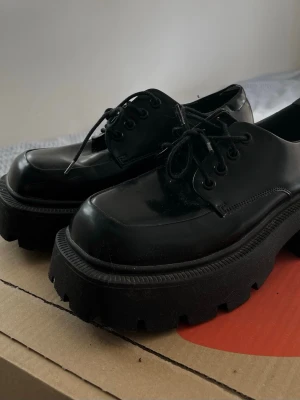 Chunky svarta loafers med platåsula - Säljer ett par svarta loafers med riktigt chunky platåsula och grovt mönstrad undersida. Skorna har snörning och är i blankt material.