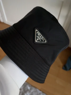 Svart Prada Hatt Bucket Hat - Snygg svart bucket hat från Prada med den ikoniska trekantiga logotypen framtill. Hatten är tillverkad i ett glansigt syntetmaterial som ger en modern och clean look. Perfekt accessoar för att lyfta din outfit.