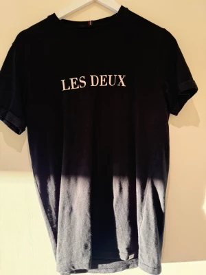 Svart t-shirt från Les Deux - Svart t-shirt från Les Deux med vit text på bröstet. Klassisk rund halsringning och korta ärmar. Tillverkad i mjuk bomull som känns skön mot huden. Perfekt för en stilren och enkel look.