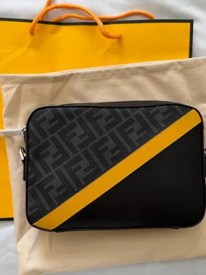 Fendi svart axelväska med gul detalj - Snygg svart axelväska från Fendi med klassiskt FF-mönster och en diagonal gul skinnrem på framsidan. Väskan har en rektangulär form och är tillverkad i canvas och läder. Perfekt för dig som gillar statement-accessoarer. Skriv dm för beställning.