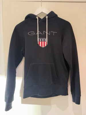 Mörkblå hoodie från GANT - Snygg mörkblå hoodie från GANT med vit broderad logga och amerikanskinspirerat sköldmärke på bröstet. Hoodien har vit dragsko i huvan och en stor magficka. Tillverkad i mjukt bomullsmaterial, perfekt för en avslappnad stil.