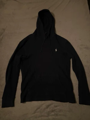Svart hoodie från Polo Ralph Lauren - Säljer en svart hoodie från Polo Ralph Lauren med klassisk huva och dragsnören. Tröjan har en liten vit broderad logga på bröstet och en stor magficka framtill. Tillverkad i mjukt, ribbat material som känns riktigt skönt mot huden.