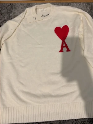 Vit stickad tröja från Ami Paris - Vit stickad tröja från Ami Paris med röd hjärtlogga och bokstaven 'A' på bröstet. Tröjan har rund halsringning och ribbade muddar vid ärmslut och nederkant. Perfekt för dig som gillar stilrena plagg med en twist.