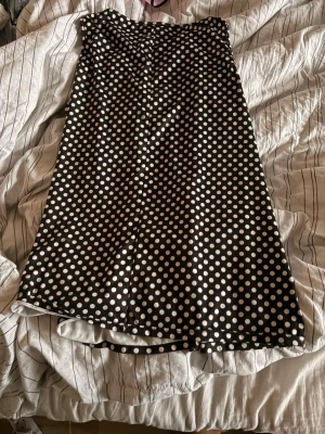 Svart prickig ärmlös topp - Svart ärmlös topp med vita prickar och dragkedja i ryggen. Toppen har en rak passform och är gjord i ett mjukt, lätt material som känns skönt mot huden. Perfekt för dig som gillar retrovibbar och vill ha ett klassiskt mönster i garderoben.