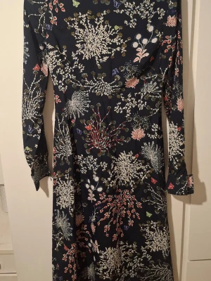 Mörkblå blommig långklänning - Snygg långklänning i mörkblått med blommigt mönster i vitt, rosa och grönt. Klänningen har lång ärm och markerad midja, vilket ger en smickrande siluett. Perfekt för dig som gillar färg och mönster! Materialet känns mjukt och följsamt.