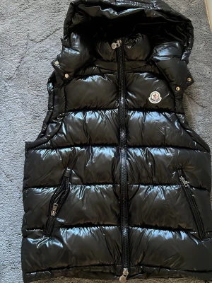 Moncler väst - Snygg svart dunväst från Moncler med glansig finish och huva. Västen har två dragkedjeförsedda fickor, Moncler-logga på bröstet och breda quiltade paneler. Perfekt för lager-på-lager och streetstyle.