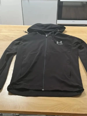 Svart hoodie från Under Armour - Svart zip-hoodie från Under Armour med huva och dragkedja framtill. Hoodien har lång ärm, två sidofickor och en liten logga på bröstet. Tillverkad i ett mjukt och stretchigt material som passar perfekt till chill eller träning. (Snöret på koftan finns ej kvar längre) 